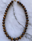 A beaded necklace made of polished brown tiger's eye beads with a small gold clasp, displayed on a white marble surface.