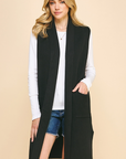 A woman modeling a long black sleeveless open-front vest over a white long-sleeve top and denim shorts, against a plain beige studio background.