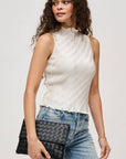 Woman wearing a white sleeveless top and blue jeans, holding a black clutch against a plain background