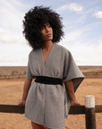 Woman wearing a gray dress with a black belt standing in a desert-like landscape.