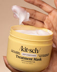 A hand holds an open yellow jar of Akit-sch Treatment Mask with coconut oil, showing white mask cream being lifted out, against a solid beige background.
