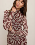 A woman wearing a long-sleeve mock-neck dress with a brown and beige zebra print pattern, posed against a solid light beige background.