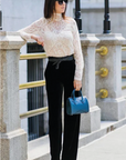 Woman in a white lace top and black pants holding a blue handbag, standing on a city street.