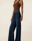 A model wearing dark blue wide-leg denim jeans and a brown ribbed tank top, standing against a plain beige studio background.
