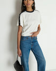 Woman wearing a white t-shirt with black sleeves and blue jeans, holding a black handbag against a plain background.