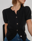 Person wearing a black cardigan with short sleeves and buttons, sitting against a plain background.