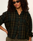 A woman wearing a dark green and black plaid button-up shirt with rolled sleeves, styled with olive green drawstring pants and black sunglasses against a plain off-white background.
