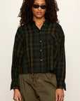A model wearing a dark green and black plaid button-down shirt with olive green drawstring pants, styled with black sunglasses against a plain off-white background.