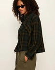 A woman with curly hair wearing a dark green and black plaid button-up shirt and olive green pants, posed against a plain off-white studio background.