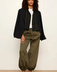A model stands against a plain off-white studio wall wearing a dark navy blazer, a green and black plaid shirt, a white top, olive green drawstring pants, and brown slip-on shoes.