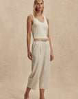 Woman wearing a white sleeveless top and beige pants against a beige background