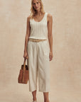 Woman wearing a white sleeveless top and beige pants, holding a brown leather bag against a beige background.