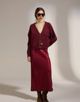 A model stands against a warm beige studio backdrop wearing a matching burgundy cardigan and midi skirt set, a cream turtleneck, layered gold necklaces, sunglasses, and brown suede boots.