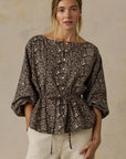 A woman wearing a dark brown and beige snakeskin print peplum blouse with three-quarter sleeves and cream pants, standing against a textured beige wall.
