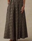 A high-waisted, full-length maxi skirt with an intricate dark brown and beige floral jacquard pattern, shown on a model wearing brown leather sandals against a textured beige studio backdrop.