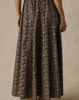 A woman wearing a long, full-length skirt with an intricate black, brown, and beige floral or lace-like pattern, paired with a black top and flat sandals, shown from the back against a neutral beige background.