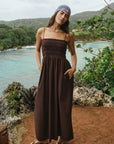 Woman in a brown dress standing by a scenic coastal area with greenery and water.