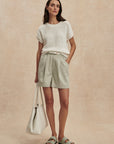 Woman wearing a white textured top and light green shorts, holding a beige bag on a beige background