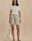 Woman wearing a white textured top and light green shorts, holding a matching tote bag on a beige background.