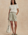 Woman wearing a white textured top and light green shorts, standing against a beige background.