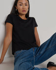 Woman wearing a black t-shirt and blue jeans sitting against a plain background
