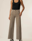 A model wearing a pair of high-waisted, wide-leg taupe jeans, paired with a black tank top and black sneakers, standing against a plain off-white studio background.