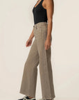A woman shown from the side wearing high-waisted, wide-leg taupe pants, a black tank top, and black-and-white sneakers against a plain off-white studio background.