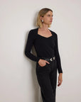 A woman with blonde hair stands against a plain light gray wall, wearing a black long-sleeve scoop-neck top tucked into black jeans with a black belt featuring silver hardware.