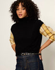 A model with curly dark hair wearing a black sleeveless mock-neck top with yellow plaid shirt sleeves, tucked into dark blue jeans with a studded belt.