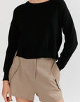 A model wearing a black long-sleeve crewneck sweater and beige shorts, cropped from the mouth down against a light gray background.