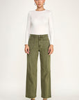 A model stands against a light gray studio background wearing a fitted white long-sleeve top, olive green wide-leg pants, and dark brown pointed-toe flats.