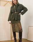 A model stands in a studio wearing an olive green utility jacket with a matching belt, a green paisley skirt, and dark brown knee-high boots.