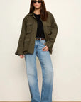 A woman modeling an olive green utility jacket over a black top and light blue wide-leg jeans, standing against a plain off-white wall.