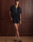 Woman in navy outfit with cap and golf shoes holding a golf club against a brown paneled wall.