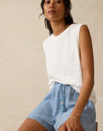 Woman wearing a white sleeveless top and blue shorts against a beige background
