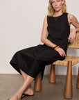 Woman in a black dress sitting on a wooden chair against a plain background