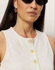 Woman wearing a white sleeveless top with yellow buttons and black sunglasses against a plain background