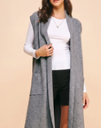 A woman models a long, sleeveless gray knit duster vest over a white long-sleeve top and black shorts, set against a solid beige studio background.