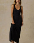 A woman modeling a black sleeveless maxi dress against a textured beige studio backdrop.
