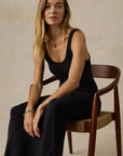 Woman in a black sleeveless dress sitting on a wooden chair against a beige background