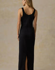 A woman wearing a sleeveless black maxi dress with a high front slit, shown from the back against a textured beige studio backdrop.
