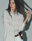 Woman wearing a light gray shirt with rolled-up sleeves, holding her hair against a plain background