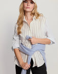 A woman modeling a light beige baseball cap, a cream and navy vertically striped button-down shirt, and a light blue knit sweater tied around her waist, paired with black pants, against a plain light gray background.