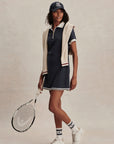 Woman in a navy and white tennis outfit holding a racket on a neutral background