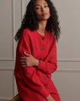 A woman with long, curly dark hair wearing a vibrant red knit cardigan dress with dark buttons, posed against a light gray paneled wall.
