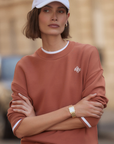 Woman wearing a brown sweatshirt with a logo, white cap, and gold watch in an urban setting.