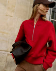 A woman wearing a bright red textured zip-up sweater, brown pants, a dark brown baseball cap, and holding a black leather bag, standing against a beige stone wall.