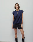 A woman modeling a navy blue short-sleeve top with a front zipper, paired with black shorts and black boots, standing against a plain light gray wall.