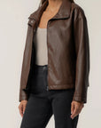 A woman wearing a brown faux-leather moto jacket over a beige tank top and black jeans, shown from the neck down against a plain light background.