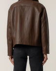 A person shown from the back wearing a cropped brown leather jacket and dark jeans against a plain light background.